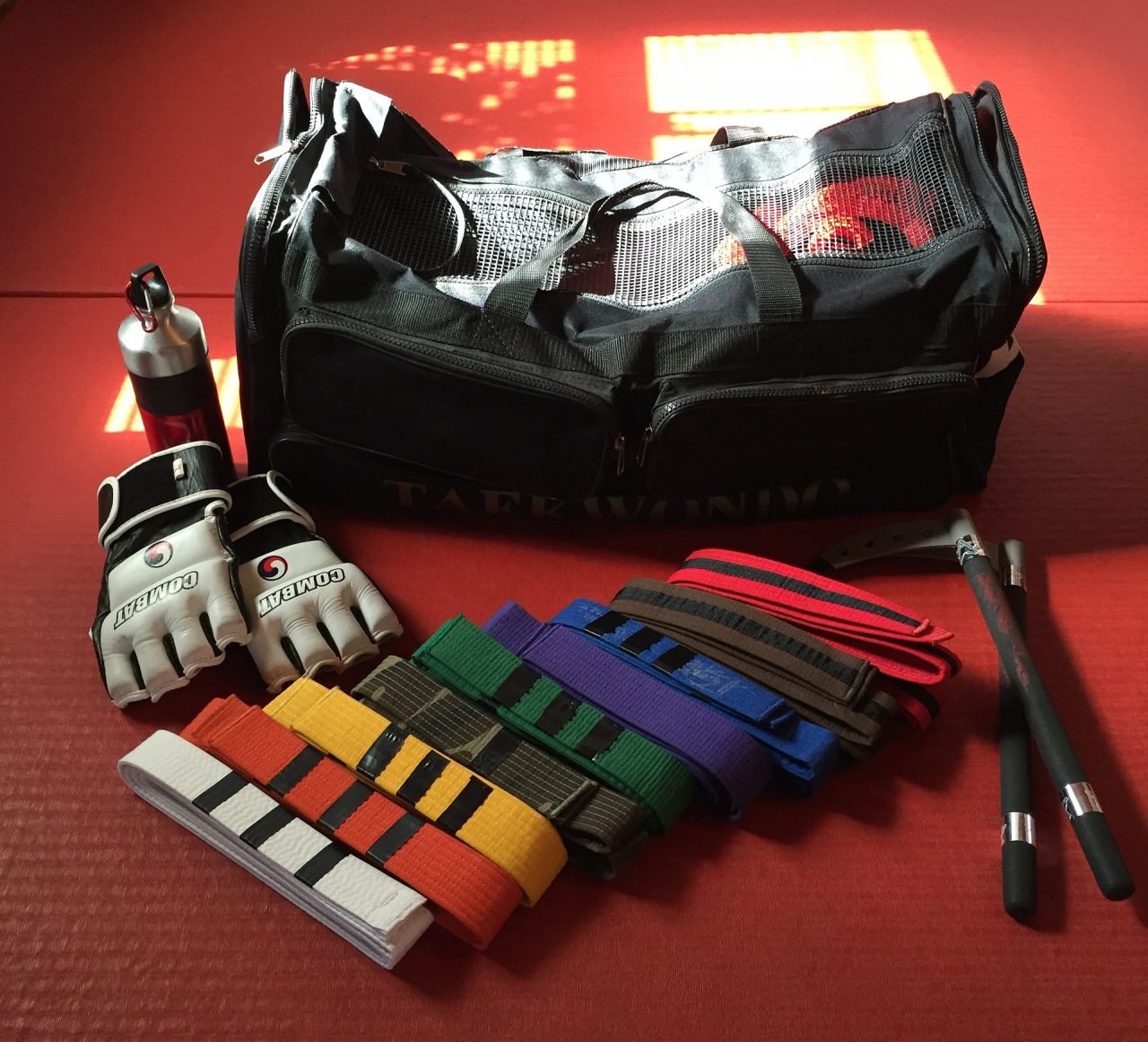Various colored martial arts belts, sparing gear, and weaponry in front of a gym bag on red carpet